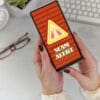 Female holding a phone at desk with Scam Alert warning screen in red and yellow