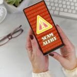 Female holding a phone at desk with Scam Alert warning screen in red and yellow